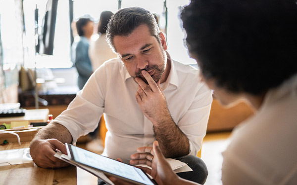 A man & woman sit together at a table, focused on a tablet, discussing B2B Commerce ROI and comparing thoughts.